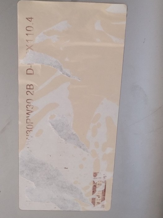Sliten etikett med delvis bortrivna delar och svaga textfragment på en ojämnt färgad bakgrund.