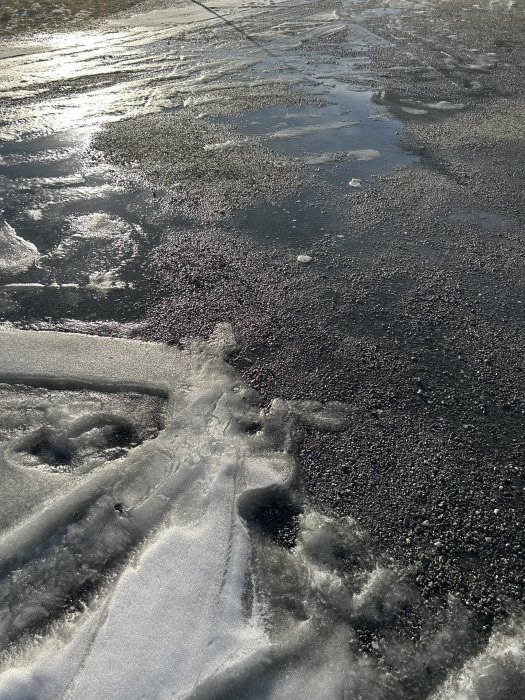 Solbelyst is och vatten på asfalterad väg; töväder; reflektioner; vinterslask; dagtid.