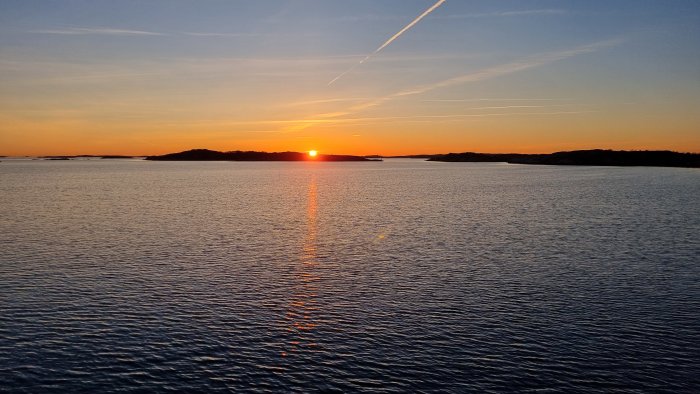 Solnedgång över havet, silhuett av öar, reflektion i vattnet, lugn, flygplansspår i himlen.