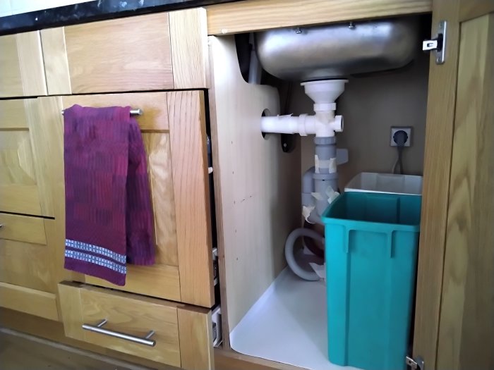 Träskåp i kök öppnat, visar vaskens rör och grönt avfallskärl. Handduk hänger på skåpsdörr.
