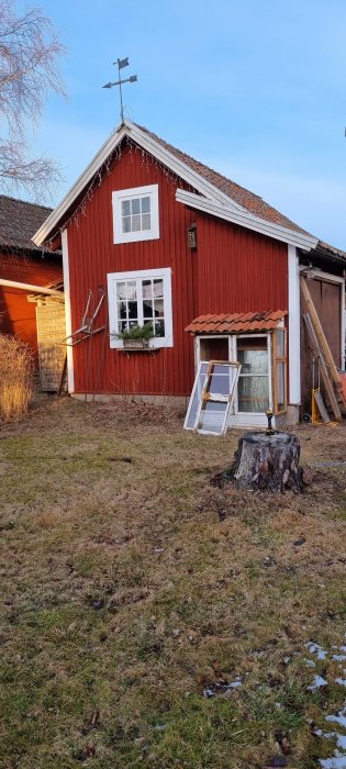 Röd stuga med vita knutar, trädstubbe, stegar, fönster, julbelysning, väderkvarn på taket, klar himmel.