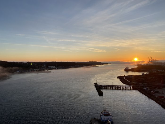 Solnedgång över hamn med fartyg, kranar och dimma längs vattenled, mot himmel med klarblå färggradient.