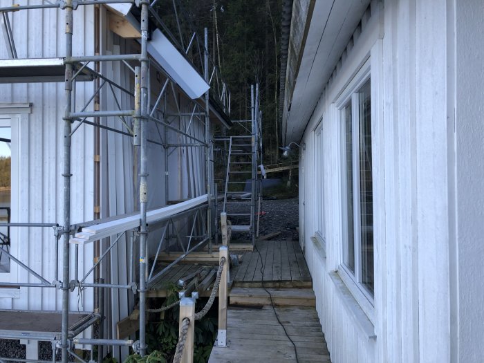 Vit byggnad under renovering med ställningar, skogsbakgrund, fönster, soligt väder, trädäck, ogjord trädgård.