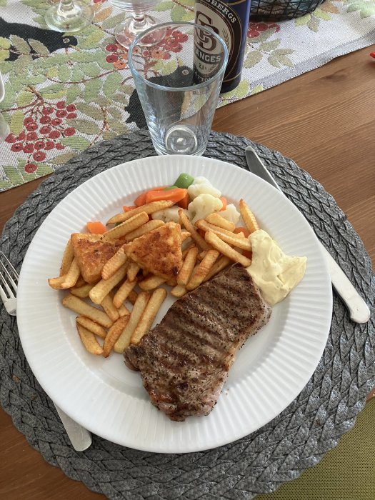 Ett måltidsfat med stekt biff, pommes frites, grönsaker och sås, ett glas vatten, ölburk i bakgrunden.