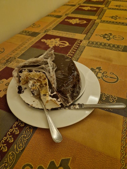 Halväten tårta med plastfolie på en vit tallrik, silverfärgad sked, färgglatt bordsunderlägg.