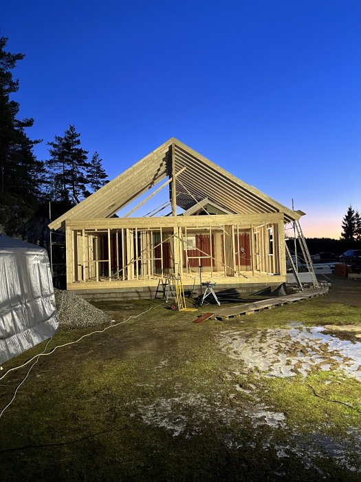 Trästomme för hus under konstruktion vid skymning med arbetsljus och lätt snötäcke på marken.