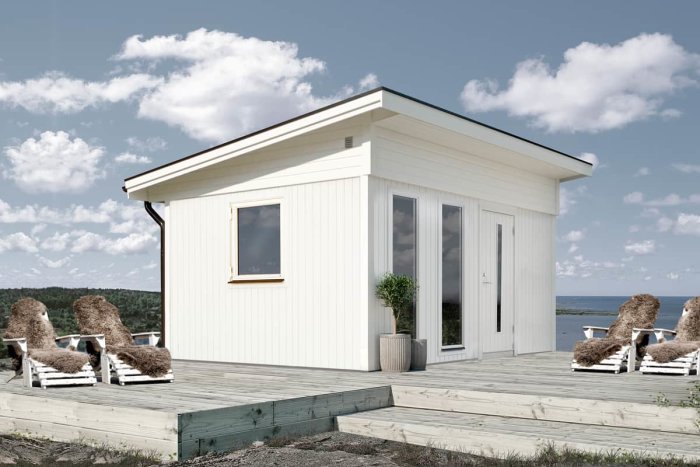 Liten vit stuga på träveranda med lounger, havsutsikt och klarblå himmel.