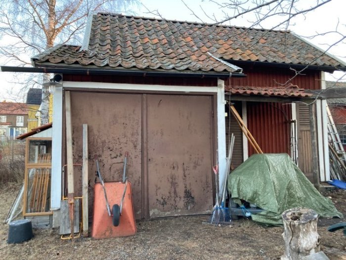 Slitet garage med rostig port, trädgårdsverktyg, presenningtäckt föremål och träbitar. Nedgången och oorganiserad uteplats.