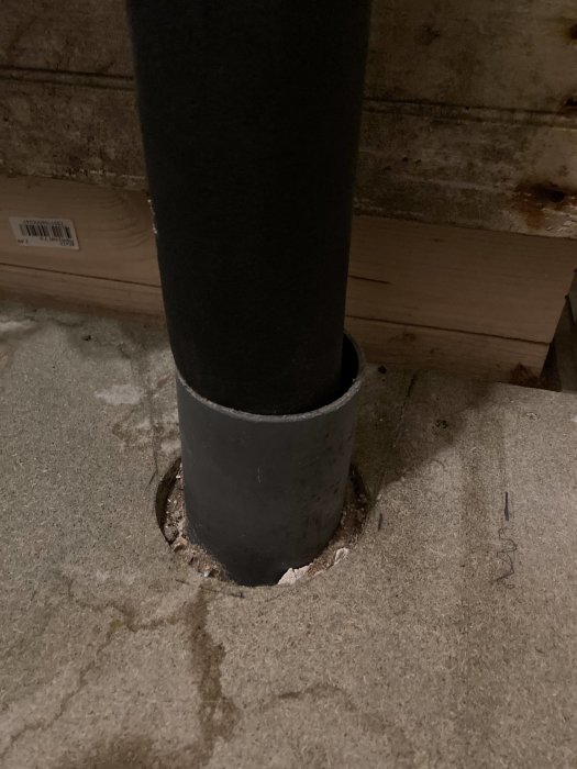Svart rör står i hål med cement, vid betongvägg och träplanka på golvet.