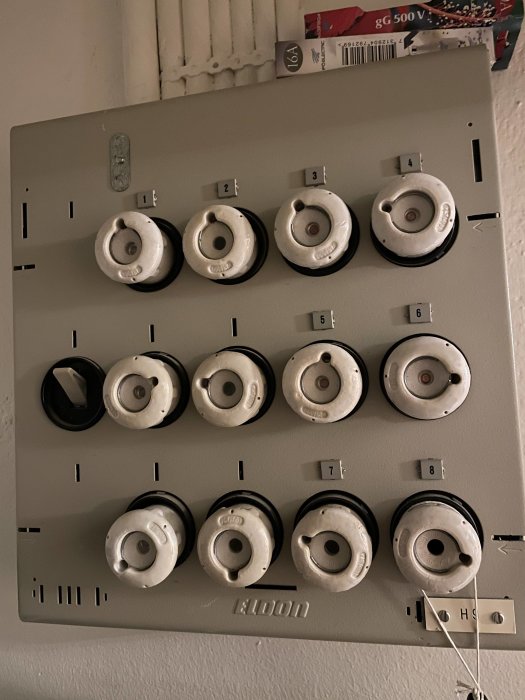 Säkringsskåp med keramiska proppar och etiketter för strömkretsar på en vägg.