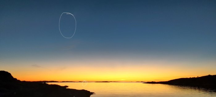 Solnedgång vid havet, färggradienter i himmeln, siluett av kustlinje, kontraster, lugnt vatten, flygplansspår.