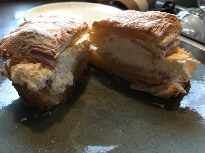 Ett bakverk, troligen en semla, delad på hälften, med vispad grädde, på grått fat.
