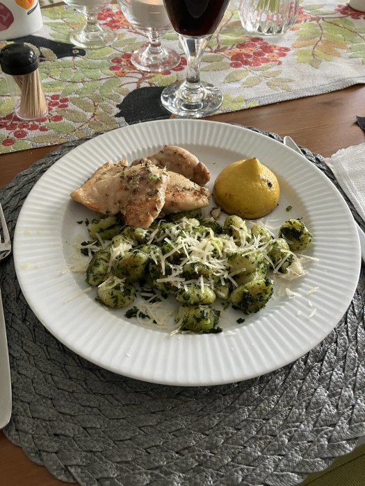 Måltid med kyckling, pestognocchi, riven ost, citron och rödvinsglas på blommig duk.