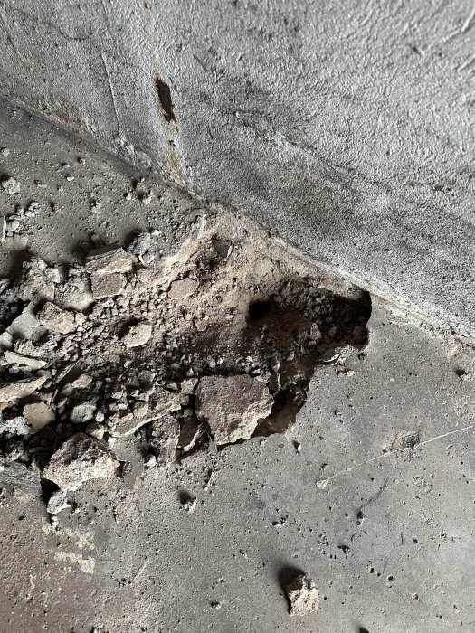 Erosion eller skada på betonggolv och vägg, exponerad armering, grus och småstenar synliga.