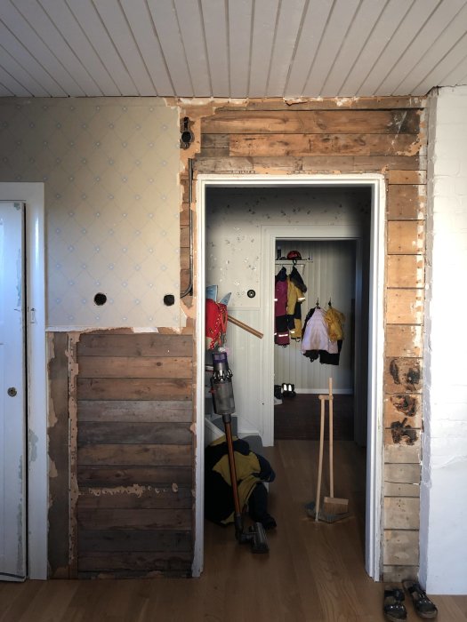 Renovering pågår, väggbeklädnad borttagen, synliga träreglar, städsaker på golvet, entré till annat rum.