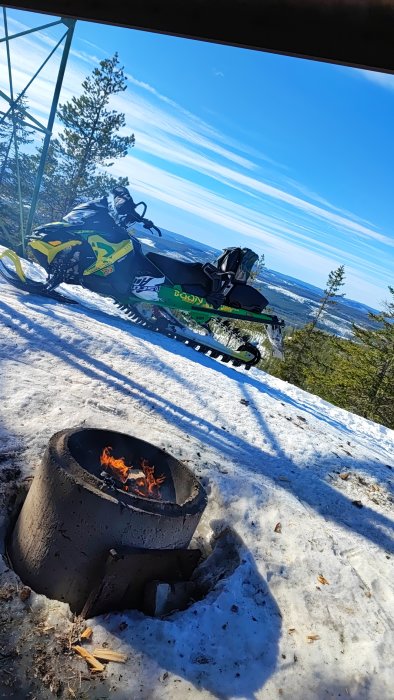 Snöskoter parkerad i snön, eld i friluftskamin, klart väder, vinterlandskap och blå himmel.