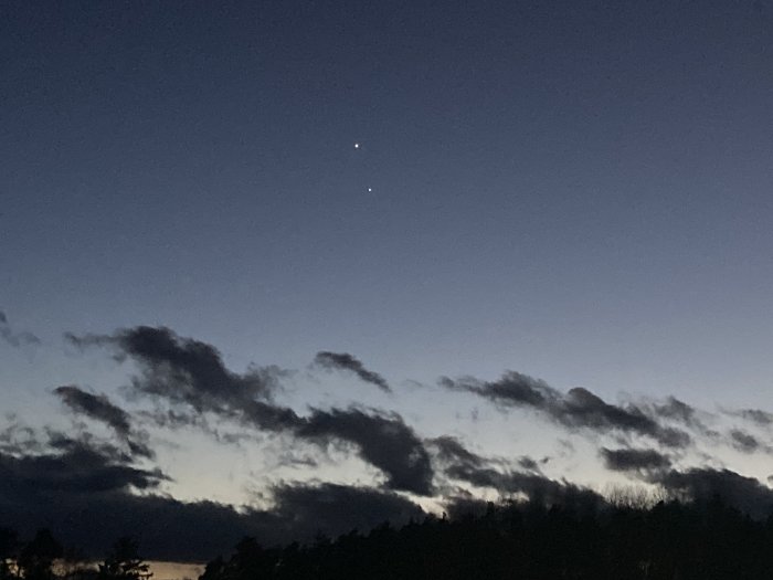 Skymning, silhuett av träd, mörka moln, två ljusa stjärnor eller planeter synliga på himlen.