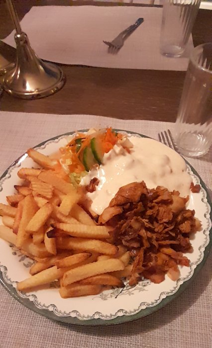 En tallrik med pommes frites, kebabkött, sallad, och sås, på ett bord med bestick och dricksglas.