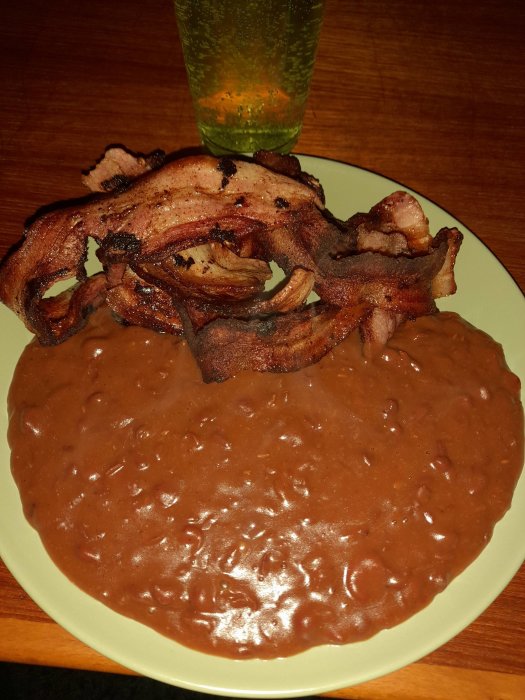 En tallrik med bönröra och bacon, ett glas med dryck, på ett träbord.