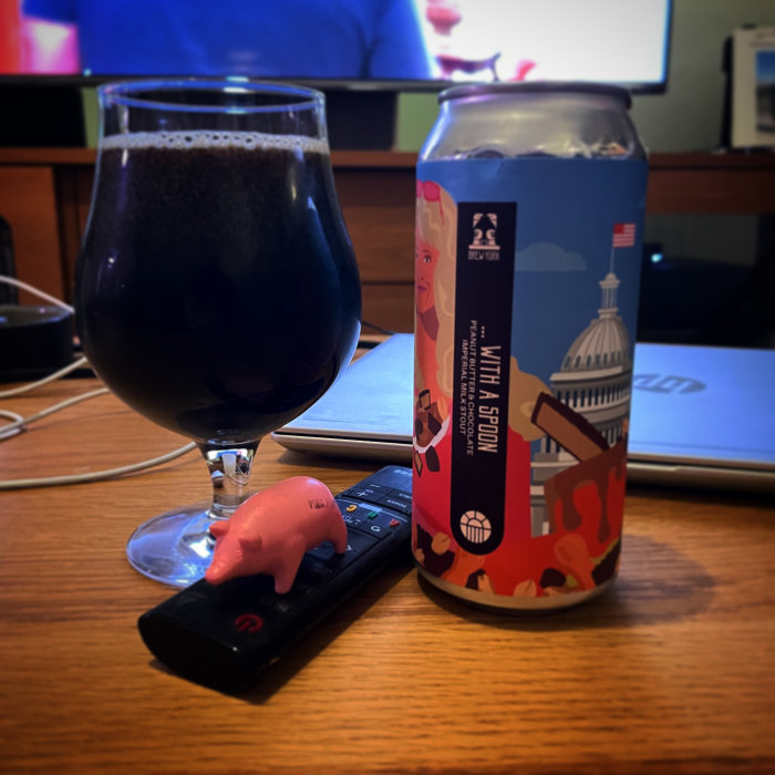 Mörkt öl i glas, ölburk, rosa grisformad pryl, fjärrkontroller, bärbar dator, suddig bakgrund med bildskärm.