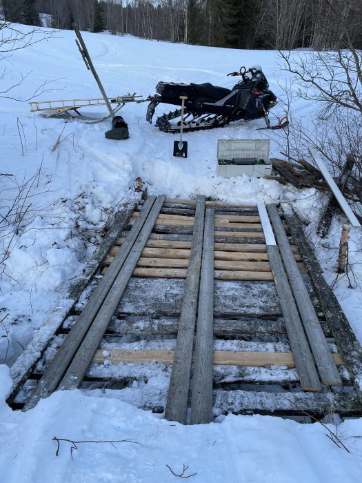 Snöskoter, släde och verktyg vid en träbro i vintertid, snö omkring och skog i bakgrunden.