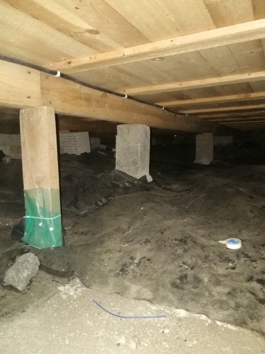 Mörkt kryputrymme under hus, träbjälkar stödda av betongpelare, synliga rör, dammigt golvyta, oisolerat.