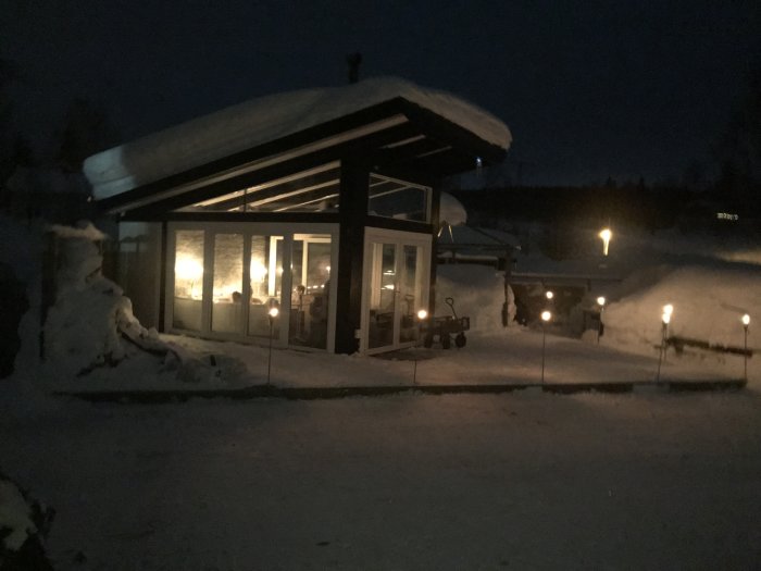 Ett hus med upplysta fönster om kvällen, omringat av snö och med tjocka snötäcken på taken.