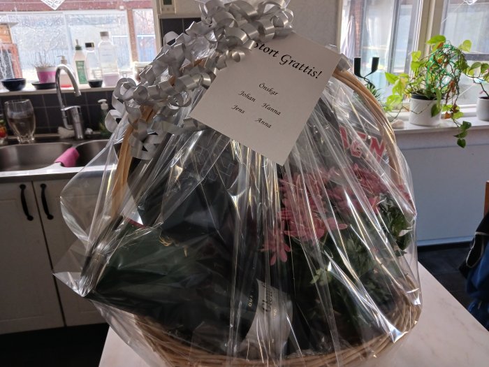 Presentkorg med cellofan, rosetter, gratulationskort, innehåller blommor, inomhus med köksbakgrund.