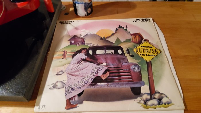 Gammal pickup, kvinna lutar sig, skylt "Nutbush City Limits", solnedgång, bergen, musikalbumomslag.