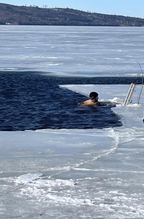 Person badar i isvak, omgiven av tjock is och snö, klar himmel i bakgrunden.