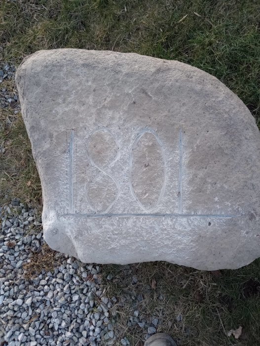 Stor grå sten med numret "1301" ingraverat ligger på marken omgiven av småsten och gräs.