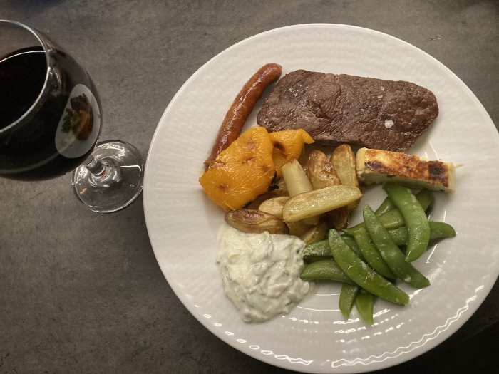 Maträtt med kött, korv, grönsaker, tzatziki på tallrik, rödvinsglas bredvid. Hyfsad presentation på mörk bakgrund.