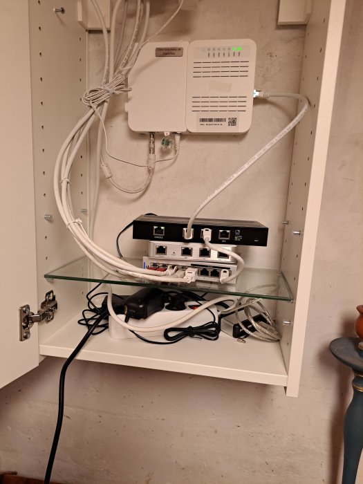 Ett nätverksskåp med router, switch, kablar och annan teknisk utrustning monterad på väggen.