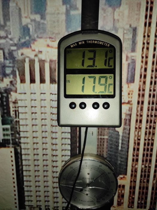 Digital termometer som visar temperaturer inomhus och utomhus, mot bakgrund av en storstadsbild; hängande analog hygrometer under.