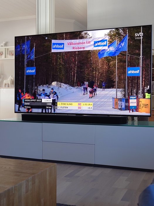 TV visar längdskidåkning, Vasaloppet 2023, skidåkare passerar blå banner, skogsbakgrund, inomhus, tävlingstid på skärmen.