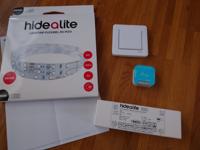 LED-strip förpackning, installationsguide, ljusdimmer och elektrisk komponent på trägolv.