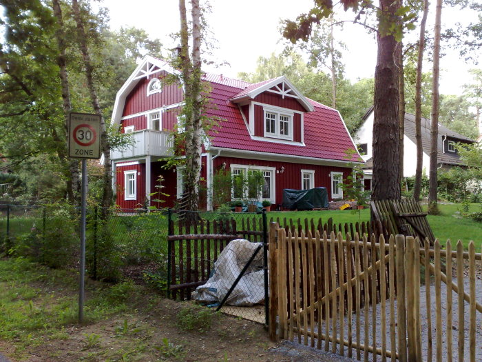 Rött hus med vita knutar, grönskande trädgård, staket, hastighetsskylt.