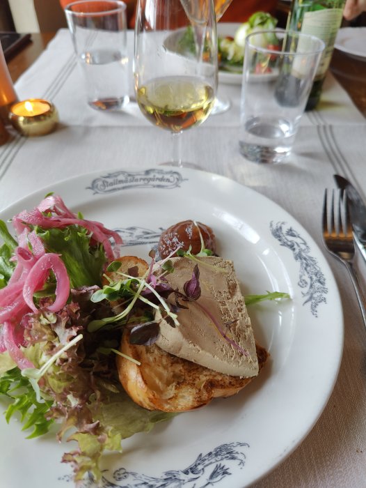 Måltid med paté på rostat bröd, sallad, picklad lök, vin, vattenglas, ljus, mysig restaurangmiljö.