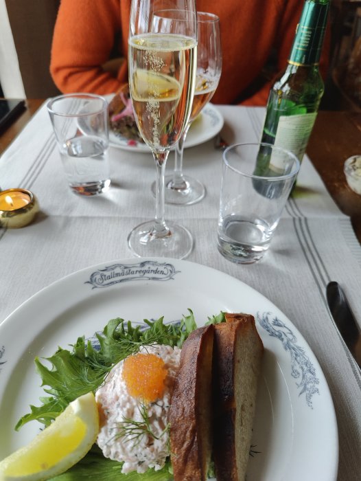 Middag med skaldjursröra, bröd, champagne, person i orange tröja, mysig atmosfär, stearinljus, vatten och ölflaska.