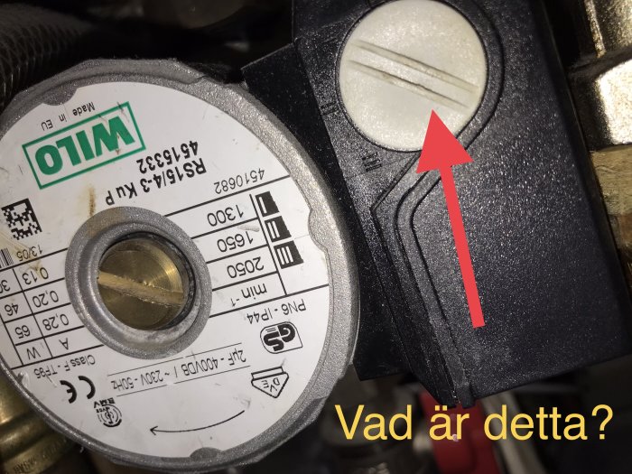 Motor- eller maskinkomponent med etikett och mätare eller knapp med röd pil och frågetext "Vad är detta?"