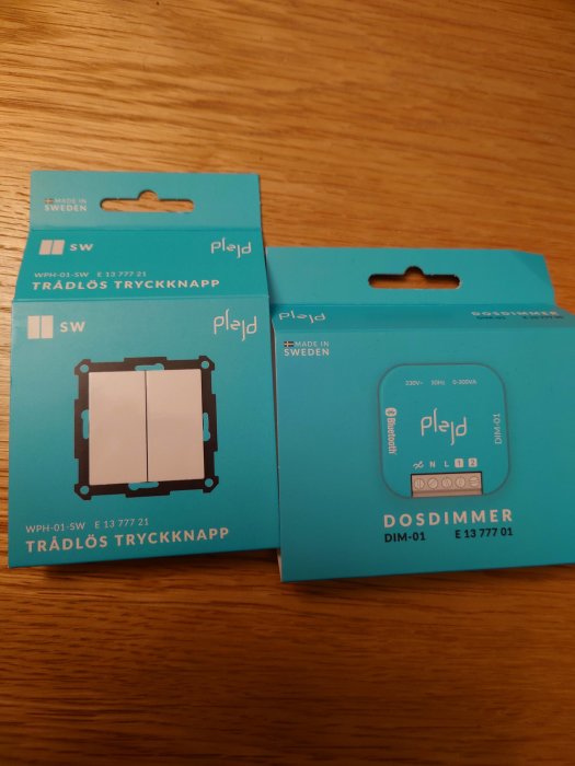 Två produkter från Plejd, trådlös tryckknapp och dosdimmer, på träbord. Förpackningar med teknisk information. Made in Sweden.