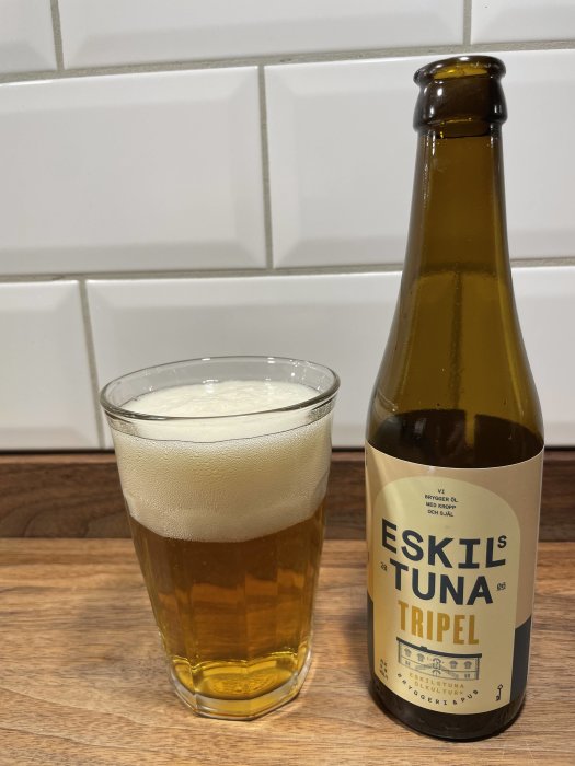 Ett glas öl och en flasketikett med "ESKILSTUNA TRIPEL". Vit kakelvägg och träyta bakgrund.