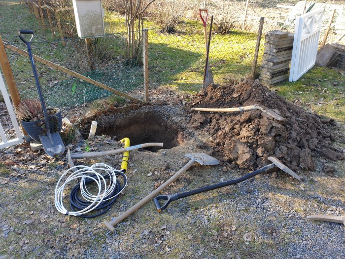 Grävd grop, jordhög, verktyg, kabel, soligt, utomhusarbete, trädgård, staket, vit grind.