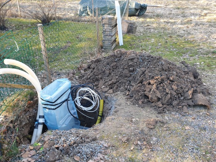 En blå behållare, slangar och kablar i en grävd grop bredvid en jordhög, utomhus vid ett staket.