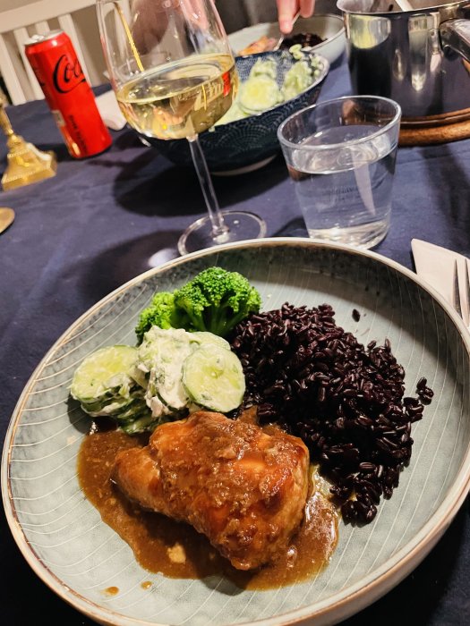 Middagstallrik med kött, svart ris, broccoli och gurksallad, glas med vitt vin och vatten, Coca-Cola burk.