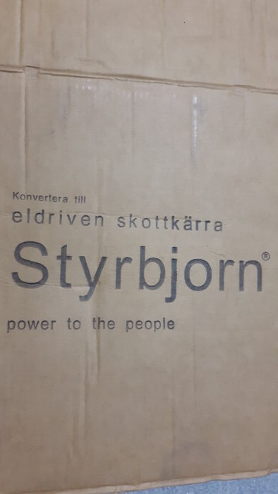 Kartong med texten "Konvertera till eldriven skottkärra Styrbjorn power to the people". Brun bakgrund, reklam eller förpackning.