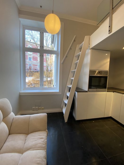 Modernt kök med vita skåp, svarta golvplattor, beige soffa, stege upp till loft, rund ljuskrona, fönster mot snö.