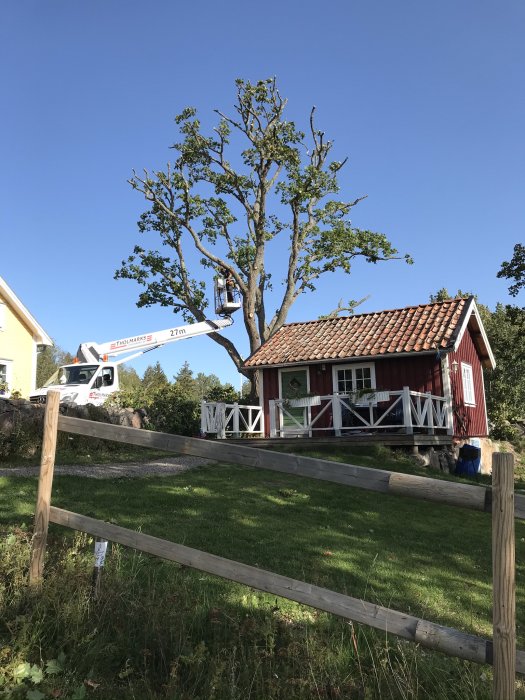 Ett rött trähus med vita knutar, trädgård, trästaket, och lift-fordon som beskär träd, klarblå himmel.