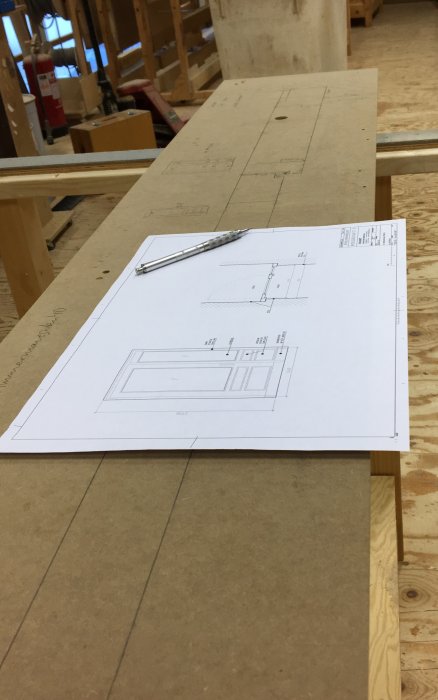 Teknisk ritning på arbetsbänk, linjal, verkstadsmiljö i bakgrunden.