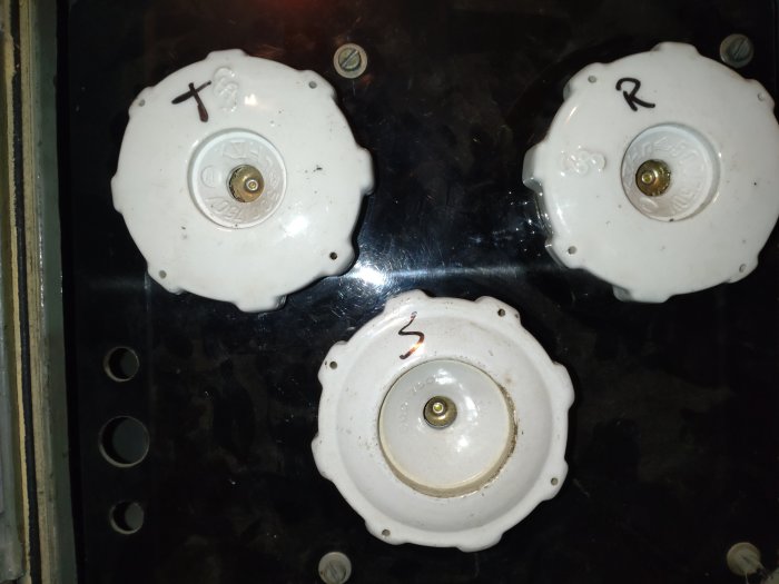 Tre vita keramiska säkringar på svart panel, märkta med X, R och S, förmodligen i ett elektriskt skåp.